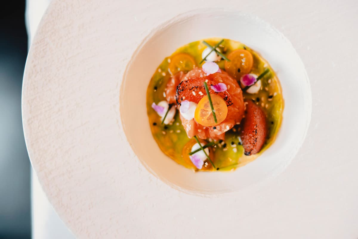 Kunstvoll angerichteter Lachs in Kräuteröl mit Kirschtomaten, essbaren Blüten und frischen Kräutern in weißer Schale.