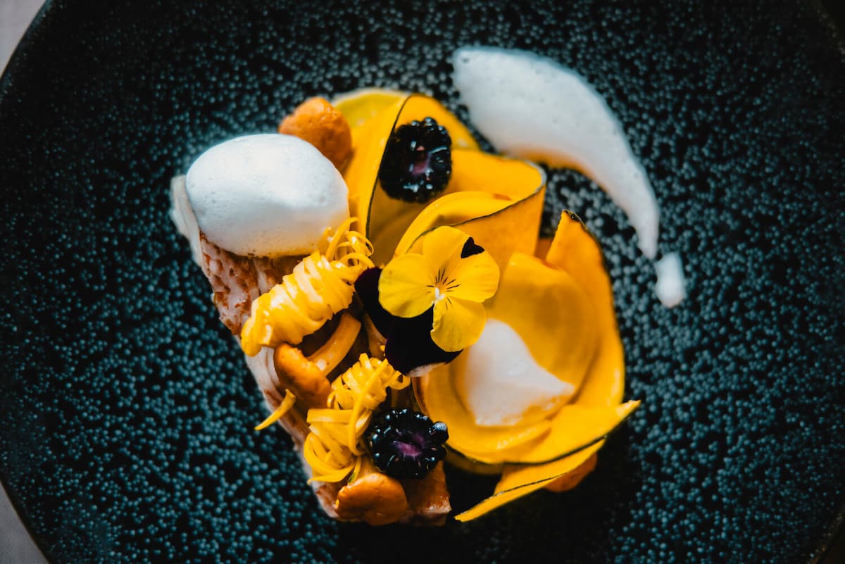 Ein kunstvoll angerichtetes Gericht auf dunklem Teller – goldene Kürbisblätter, leichter Schaum, essbare Blüten und Beeren in leuchtendem Farb- und Texturspiel.