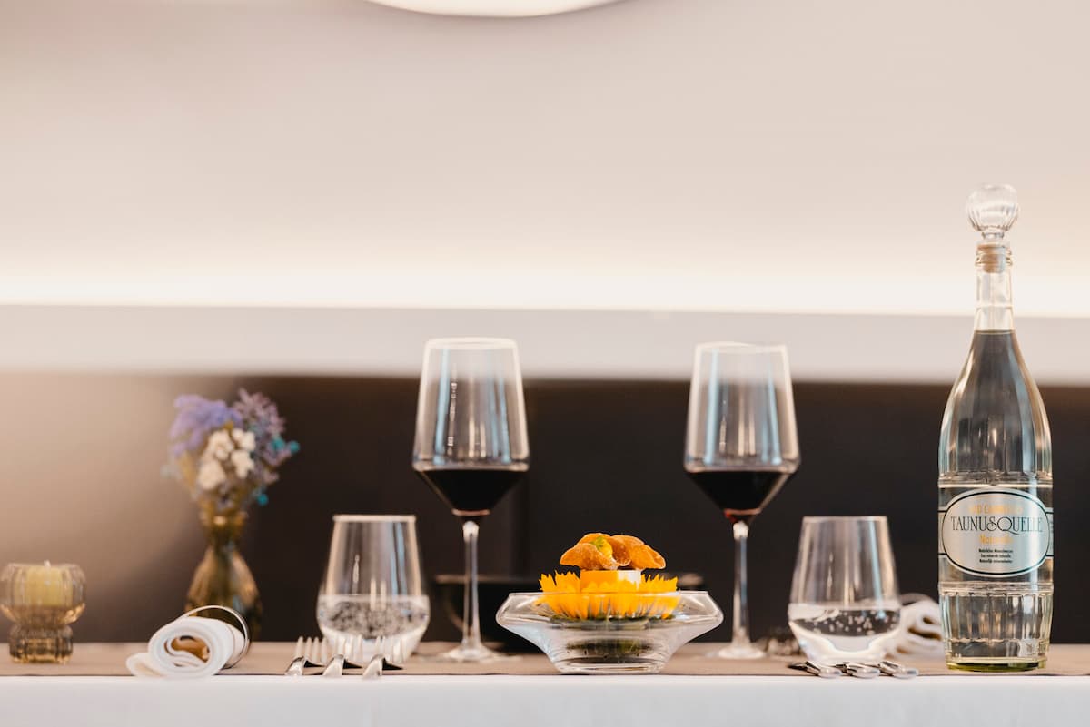 Ein elegant gedeckter Restauranttisch mit Weingläsern, Wasser und einem Gericht mit gelben Blütenblättern, sanft beleuchtet vor schlichtem Hintergrund und kleiner Blumenvase.
